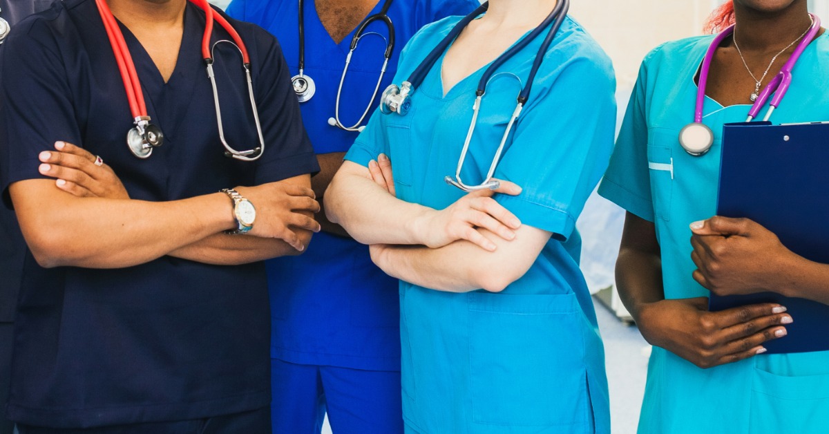 Medical team in blue and green scrubs, stethoscopes visible, standing together with folded arms and documents.