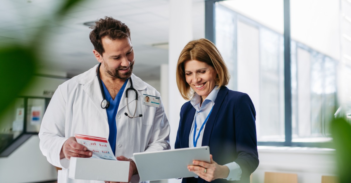 A healthcare worker and colleague smile over a tablet and paperwork in a sunlit clinic office with large windows.