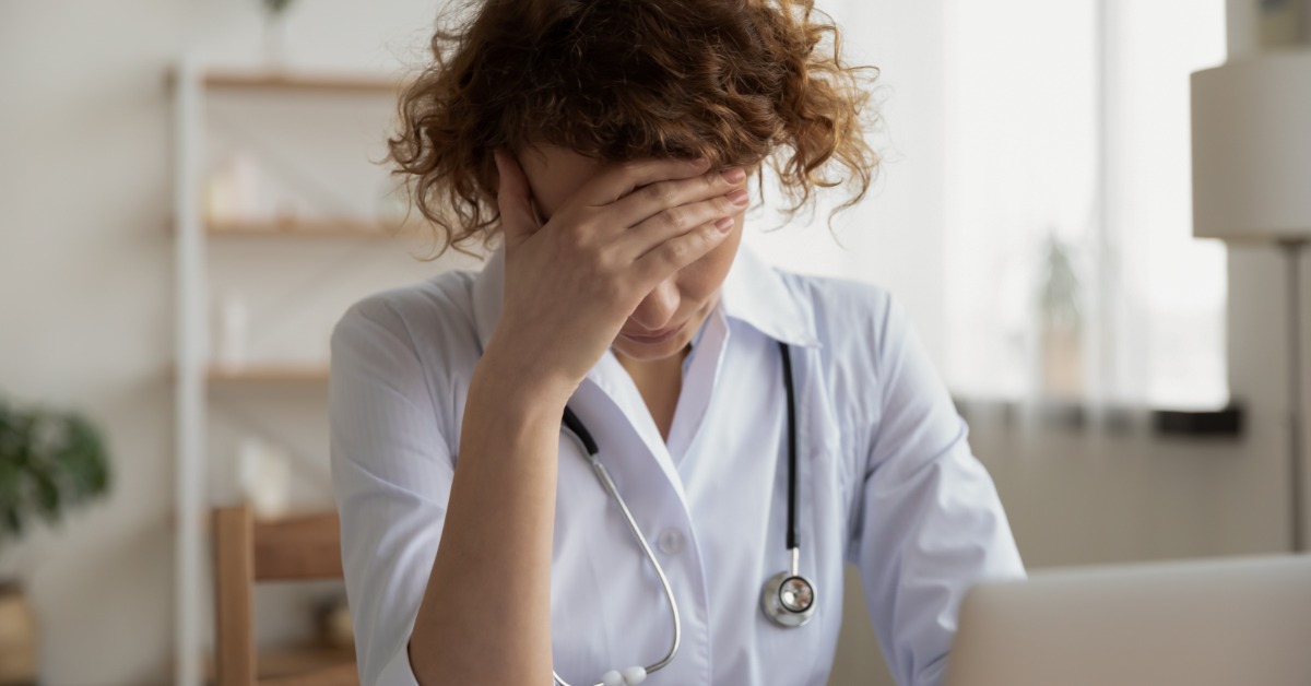 A clinician in a white coat rests one hand over their face beside a laptop, with a stethoscope around their neck.