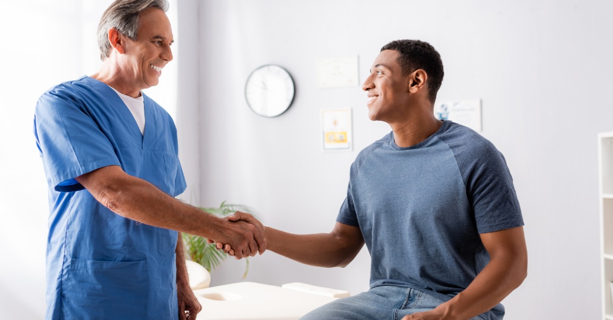 Smiling patient and healthcare professional shake hands during a clinic visit, facing each other beside an exam table.