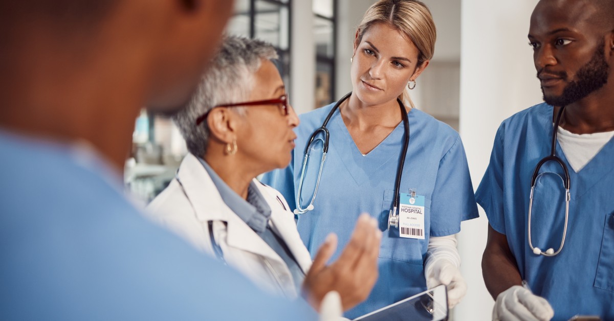 A group of medical professionals stands around a doctor wearing a white lab coat. The doctor is speaking to them.